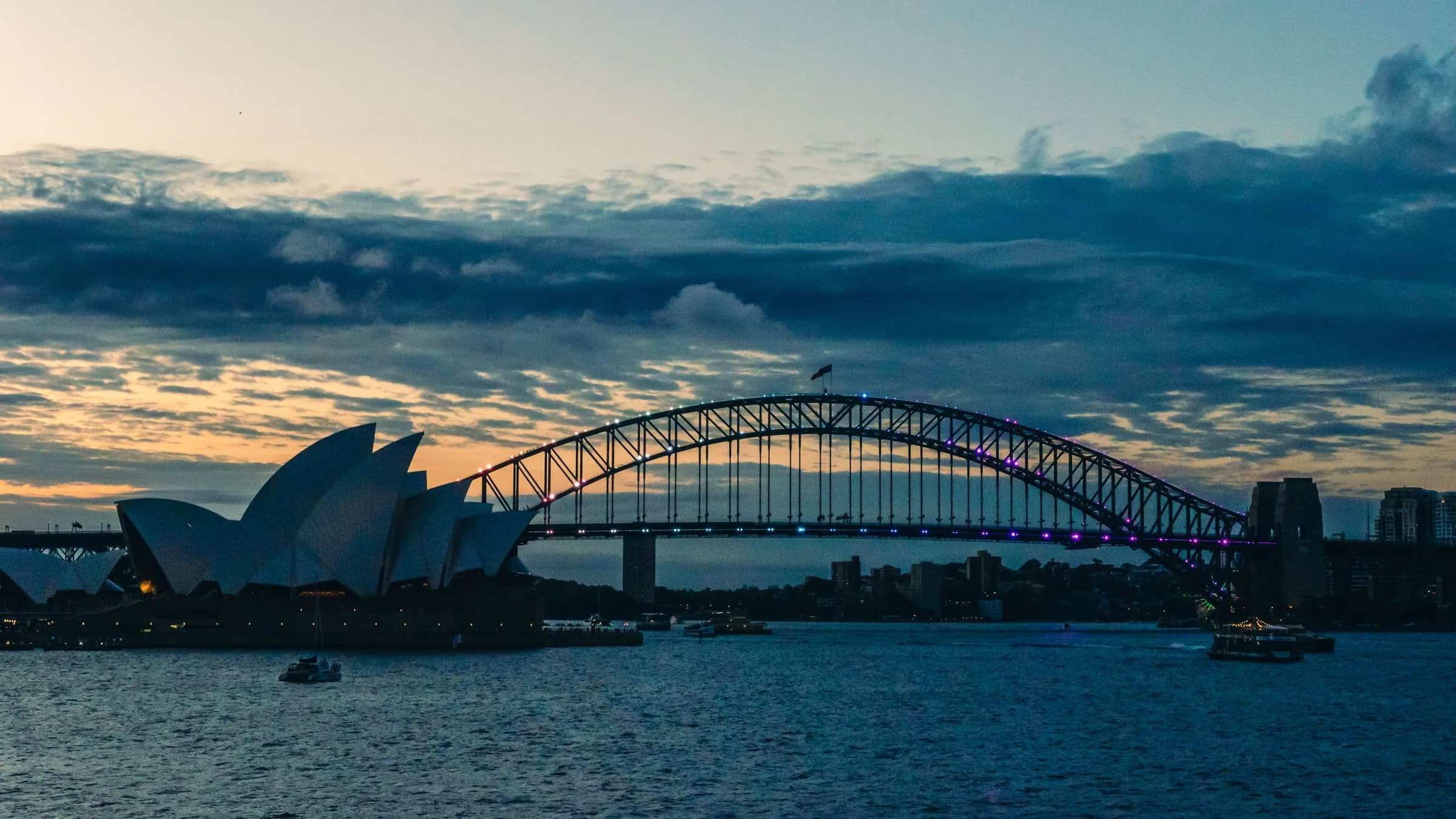 Sydney Harbour at dusk, with the Opera House and Harbour Bridge lit against the city skyline